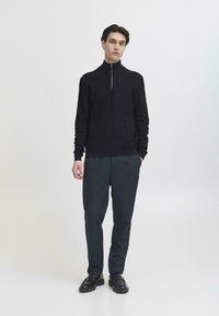 Marineblaues Zip-Up-Sweatshirt mit strukturiertem Strickmuster, stehendes Modell in dunklen Hosen und schwarzen Schuhen vor einfarbigem Hintergrund.
