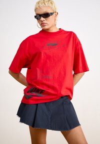 Raudona per didelė marškinė su juodu Nike logotipu, derinama su tamsiai mėlynu klostuotu sijonu. Pasižymi trumpomis rankovėmis ir laisvu kirpimu.