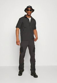 Crna polo majica kratkih rukava s patentnim zatvaračem, uparena s tamnim cargo hlačama koje imaju bočne džepove i kontrastne zakrpe. Crni bucket šešir i sunčane naočale.