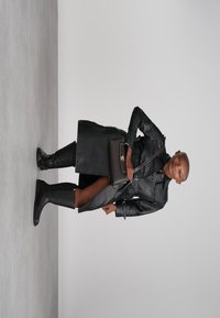 Person in black leather trench coat, knee-high black boots, and carrying a black purse, standing against a plain light gray background.