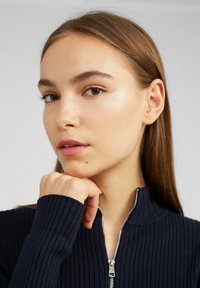 Marineblauer Ripp-Pullover mit einem Halb-Reißverschluss-Kragen, ausgestattet mit metallischen Reißverschlussbeschlägen und einem strukturierten Stoff. Die Hand des Modells ruht auf dem Kinn.