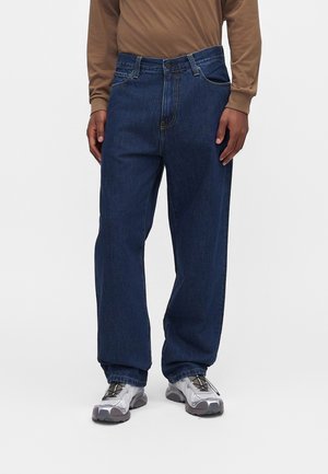 Person iført mørkeblå løse jeans, beige langærmet skjorte og grå sportssko, der står på en ensfarvet lys baggrund.