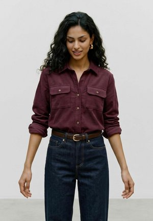 Femme aux longs cheveux bouclés et foncés, portant une chemise bordeaux boutonnée avec les manches retroussées et un jean sombre avec une ceinture marron, regardant vers le bas et souriant.