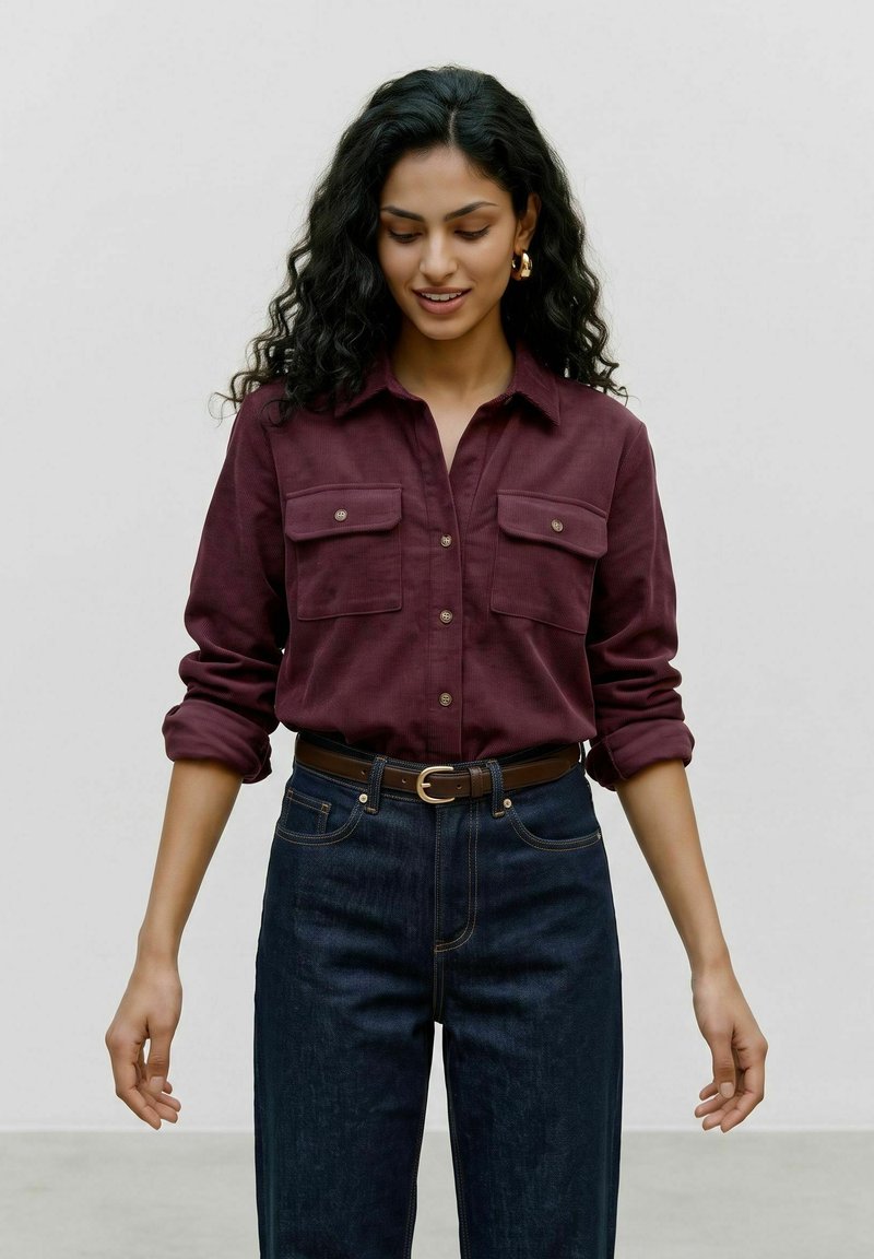 Femme aux longs cheveux bouclés et foncés, portant une chemise bordeaux boutonnée avec les manches retroussées et un jean sombre avec une ceinture marron, regardant vers le bas et souriant.
