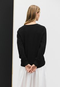 Maglione nero a maniche lunghe con scollatura rotonda. Tessuto morbido, vestibilità comoda, polsini a costine. Le mani del modello sono incrociate dietro la schiena, saio bianco visibile.