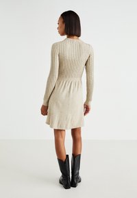 Robe en tricot beige avec texture côtelée, corsage ajusté, jupe évasée et manches longues, associée à des bottes noires montantes.