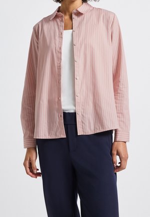 Woman wearing a light pink, vertical striped button-up shirt over a white top and dark navy pants, standing against a plain background.