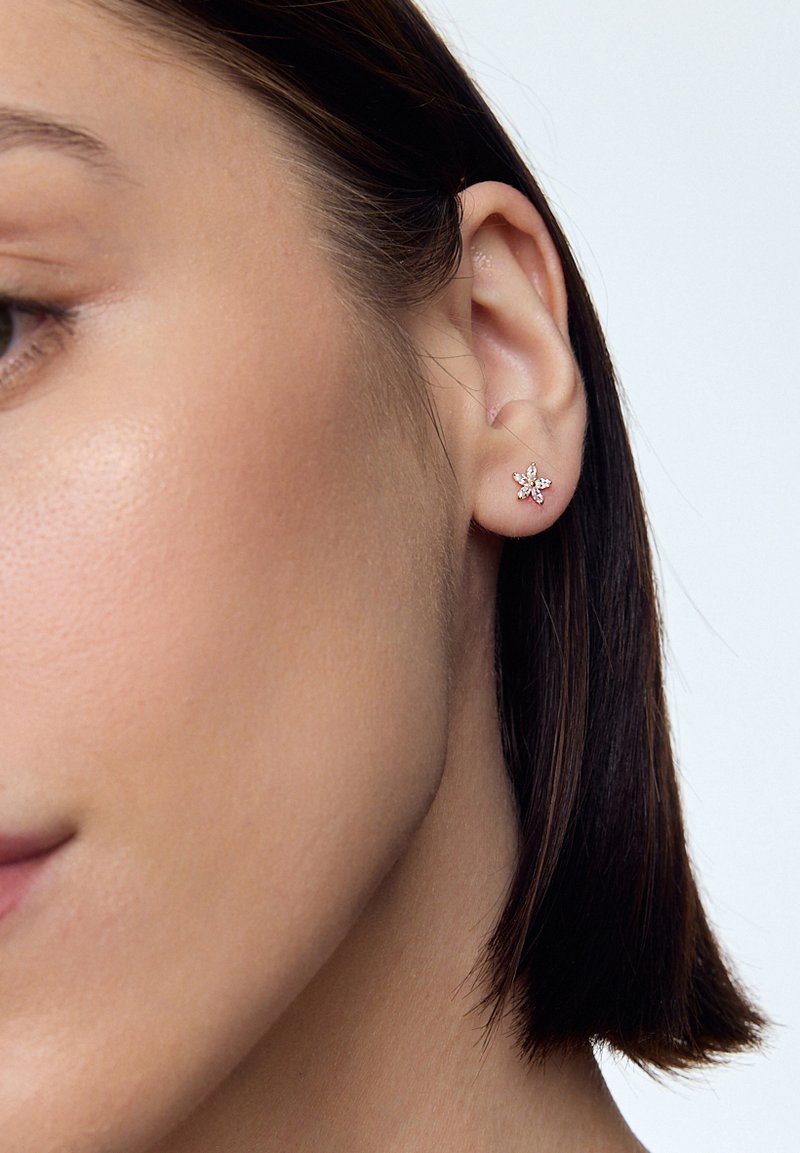 Złota kolczyk w kształcie kwiatu, z małymi, przezroczystymi kamieniami jako płatkami, wyeksponowany na ciemnym tle włosów.