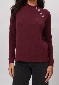 Pull en maille bordeaux avec une texture côtelée, col montant et quatre boutons décoratifs sur l'épaule gauche. Associé à un pantalon noir.