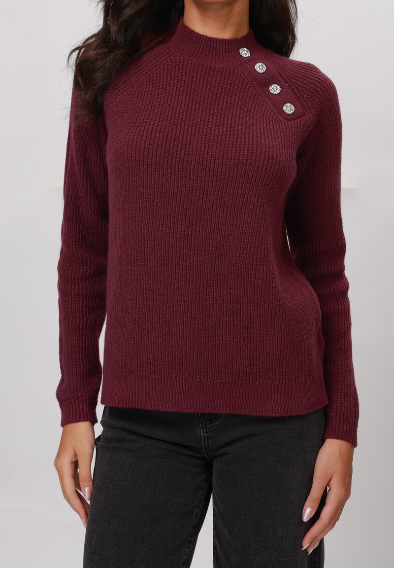 Pull en maille bordeaux avec une texture côtelée, col montant et quatre boutons décoratifs sur l'épaule gauche. Associé à un pantalon noir.
