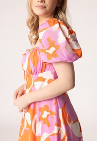 Roze en oranje bloemenjurk met pofmouwen, geplooide halslijn en vloeiende stof. Kenmerkt zich door gedurfde kleuren en grote, abstracte patronen.