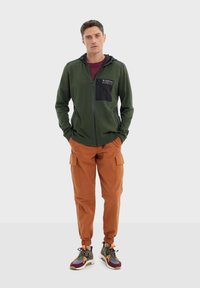 Veste à capuche zippée verte avec poche poitrine noire, pantalon cargo orange avec poignets élastiques, et chaussures de sport multicolores. Texture du tissu lisse.