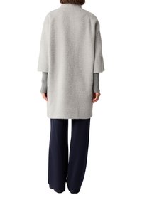 Persona in piedi, che indossa un cappotto a metà lunghezza grigio chiaro con texture sopra una maglia a maniche lunghe grigia a coste e pantaloni larghi blu navy scuri, di spalle.