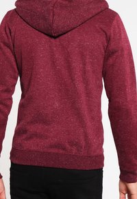 Bordeauxrode capuchontrui met een gestructureerde stof, lange mouwen en een geribbelde zoom. Voorzien van een capuchon met trekkoorden en geen zichtbare logo's.