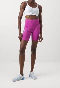 Růžové vysoké sportovní šortky z elastického materiálu, kombinované s bílým sportovním podprsenkou s černými akcenty. Na nohou jsou bílé a modré sportovní boty.