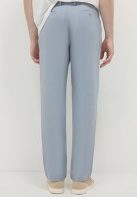 Vista posteriore di una persona che indossa pantaloni su misura blu chiaro, un top bianco e scarpe beige, in piedi contro uno sfondo bianco puro.