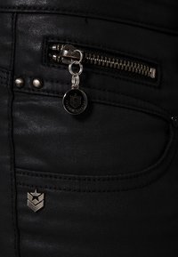 Jeans de mezclilla negra con un bolsillo con cremallera y herrajes plateados, un llavero decorativo y un pequeño logo cerca del bolsillo.