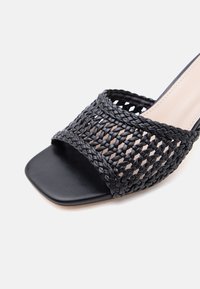 Mule noire à bout ouvert avec un bout carré et une large lanière en cuir tressé tissé sur le devant.