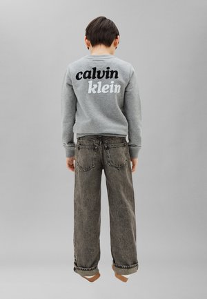 Barn stående barfodet med ryggen mod kameraet, iført en grå Calvin Klein sweatshirt og løstsiddende mørke jeans mod en ensfarvet baggrund.