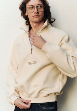 Cremfarbener Quarter-Zip-Pullover aus weichem Stoff, mit einer Fronttasche, gerippten Bündchen und einem gestickten Logo auf der Brust.