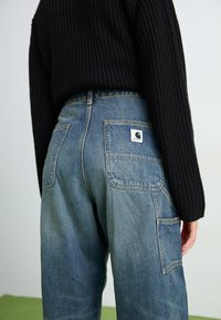 Pantaloni cargo in denim blu con due tasche posteriori e una patch con logo. La vita presenta una leggera curvatura, abbinata a un maglione in maglia nera testurizzata.