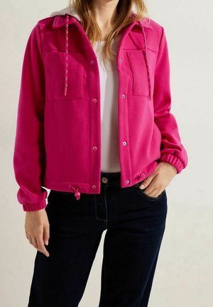 Femme portant une veste rose vif boutonnée avec cordons, poignets élastiques, chemise blanche et jean foncé, main dans la poche.