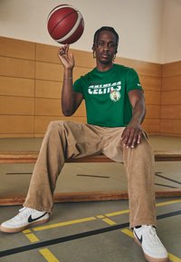 Zöld Boston Celtics póló, bézs kordbársony nadrág és fekete részletekkel ellátott fehér sportcipő. Kosárlabda egyensúlyozva az ujjak hegyén, semleges edzőterem környezet.