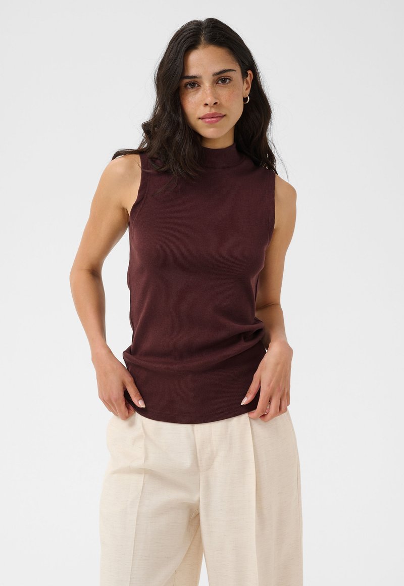 Femme aux cheveux bouclés foncés portant un haut marron foncé sans manches et un pantalon large beige clair, debout les mains près des poches.