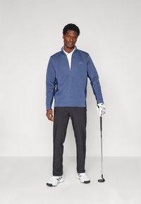 Pullover da golf blu con chiusura zip e tessuto texturizzato, abbinato a pantaloni neri e scarpe da golf bianche. Il soggetto tiene un bastone da golf, in piedi su uno sfondo neutro.