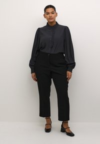 Černá blůza s knoflíky a volány, spárovaná s černými kalhotami s vysokým pasem. Modelka nosí černé boty na vysokém podpatku s pásky kolem kotníků.