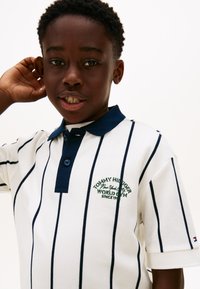 Λευκό πόλο μπλουζάκι με μπλε-σκούρες ρίγες και γιακά, με πράσινο κεντημένο λογότυπο και κουμπιά. Το ύφασμα φαίνεται υφασμένο, με κοντά μανίκια.