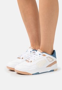 Baskets blanches avec tige en cuir, accents bleu clair et détails beige. Design perforé sur l'avant, lacets plats, et semelle en caoutchouc.