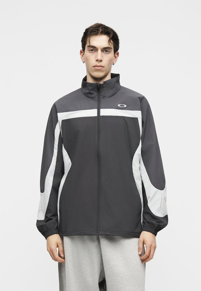 Joven vestido con una chaqueta deportiva gris y blanca con cremallera y pantalones deportivos grises, de pie contra un fondo claro y liso.