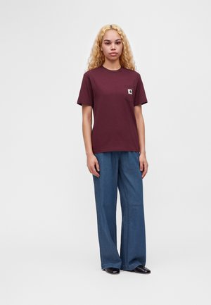 Femme aux longs cheveux blonds bouclés portant un t-shirt bordeaux à manches courtes et un jean bleu à jambes larges, debout devant un fond blanc uni.