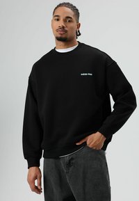 Sweat-shirt noir avec poignets et ourlet côtelés, arborant un texte brodé "RAÍCES VIVAS" en bleu clair sur la poitrine, associé à un pantalon gris foncé.