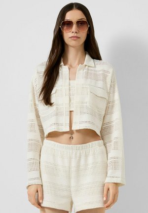 Vrouw met lang donker haar die grote zonnebrillen draagt, een wit gehaakt cropped jasje, bijpassende shorts en een zichtbare navelpiercing.