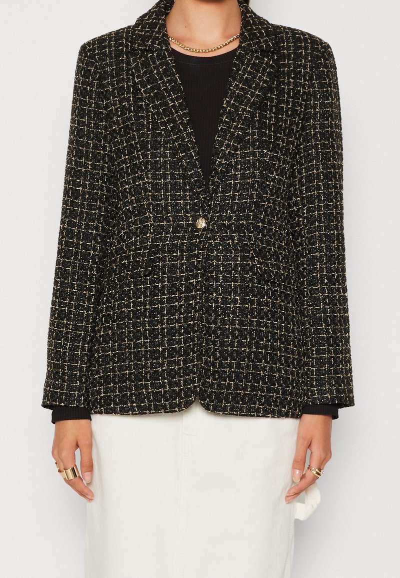 Femme portant un blazer en tweed noir et or sur un haut noir, une jupe blanche, un collier chaîne en or et des bagues en or aux deux mains.