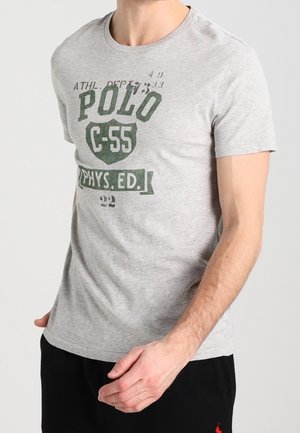 Man draagt een lichtgrijs T-shirt met korte mouwen waarop groene tekst staat: "POLO C-55 PHYS. ED." en zwarte broek.