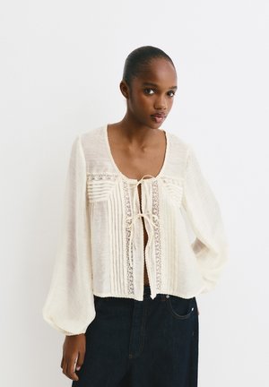 Helle Cremebluse mit langen, puffigen Ärmeln, rundem Ausschnitt, vorderer Bindegarnitur und vertikalen Falten mit Spitzenakzenten.