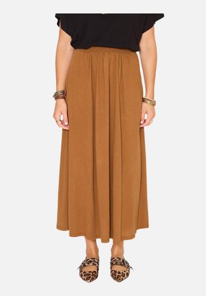 Femme portant une longue jupe marron, un haut noir, des chaussures à motif léopard avec des boucles, et plusieurs bracelets aux deux poignets sur un fond uni.