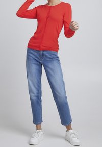 Chaqueta de punto roja de manga larga con botones, que presenta un frente con borde festoneado, combinada con jeans de mezclilla rectos de color azul claro y zapatillas blancas.
