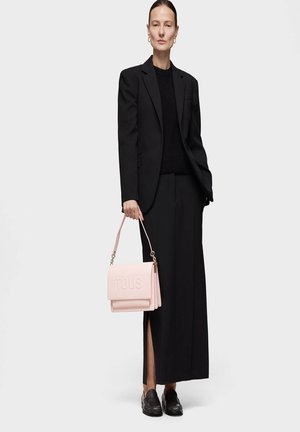 Blazer noir ajusté et pantalon assorti associés à un pull noir texturé. Un sac à main en cuir rose avec le logo "TOUS" embossé et des accents dorés.