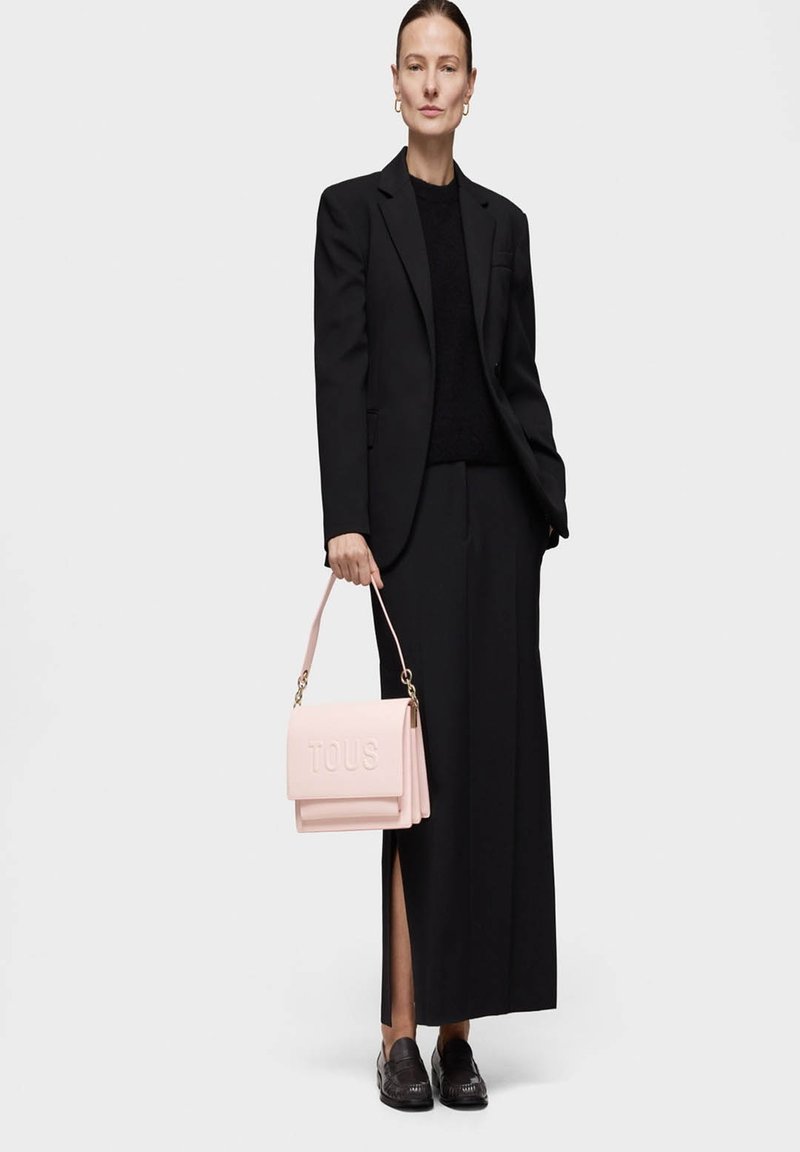 Schwarzer maßgeschneiderter Blazer und passende Hose, kombiniert mit einem strukturisierten schwarzen Pullover. Eine rosa Lederhandtasche mit geprägtem "TOUS"-Logo und goldenen Akzenten.