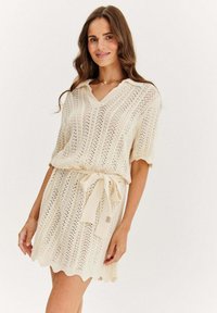 Sukienka dzianinowa w kolorze kremowym z dekoltem w kształcie litery V i krótkimi rękawami. Posiada paskowane wcięcie w talii oraz falbaniasty dół. Całość ozdobiona wzorem z otwartych splotów.