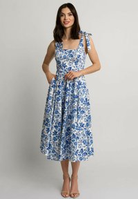 Niebieska i biała sukienka w kwiaty z kwadratowym dekoltem, związanymi ramiączkami i kieszeniami. Tkanina jest gładka i ma klasyczny wzór kwiatowy.