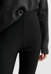 Leggings noires taille haute avec une texture côtelée, présentées à côté d'un pull en maille gris foncé. Mise en avant du tissu lisse et de la coupe ajustée.