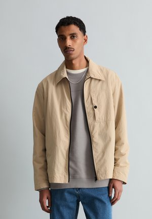 Ung mand med kort krøllet hår iført en beige lynlåstrøje over en grå sweatshirt og blå jeans, stående foran en ensfarvet grå baggrund.