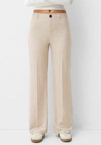 Beiga vida byxor i ett slätt tyg, med knappstängning, bälteshällor och framveck. Accenterade med vita sneakers.