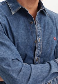 Camicia di jeans di colore blu medio con maniche lunghe, colletto button-down, cuciture a contrasto e un piccolo logo rosso sul lato sinistro del petto.
