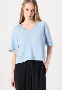 Top azzurro chiaro a V, con maniche corte, realizzato in tessuto morbido, caratterizzato da una vestibilità comoda e un orlo leggermente cropped, abbinato a pantaloni neri.